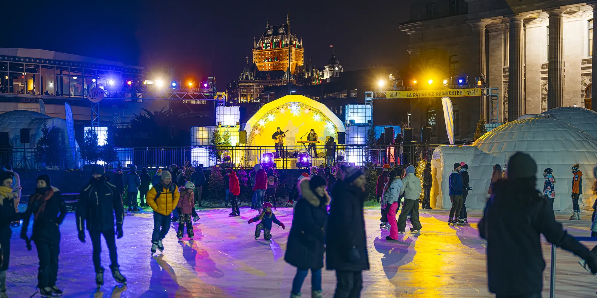 Skaters on a lit-up outdoor ice rink, with a concert in the background.
