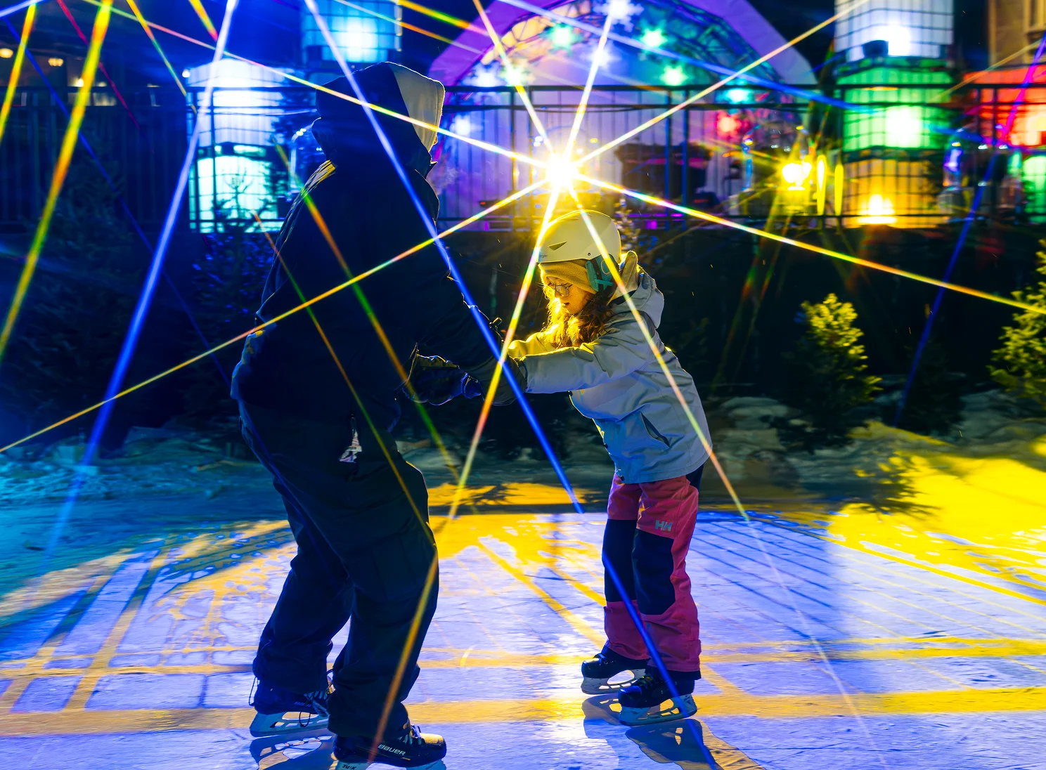 Child and adult ice skating under colorful lights at night.
